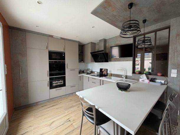 Cuisine moderne avec armoires intégrées beige, électroménagers encastrés, îlot central blanc avec tabourets, sol en bois clair et luminaires suspendus noirs, offrant un espace fonctionnel et design.