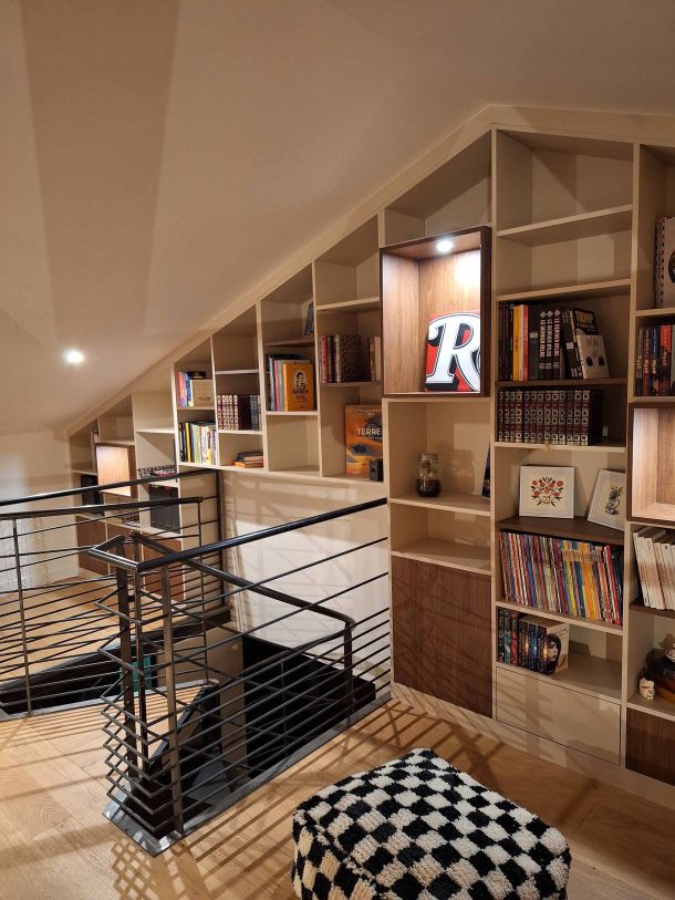 Bibliothèque sur mesure sous pente avec étagères ouvertes et niches éclairées, intégrée dans un espace en mezzanine avec garde-corps en métal noir et sol en parquet clair.